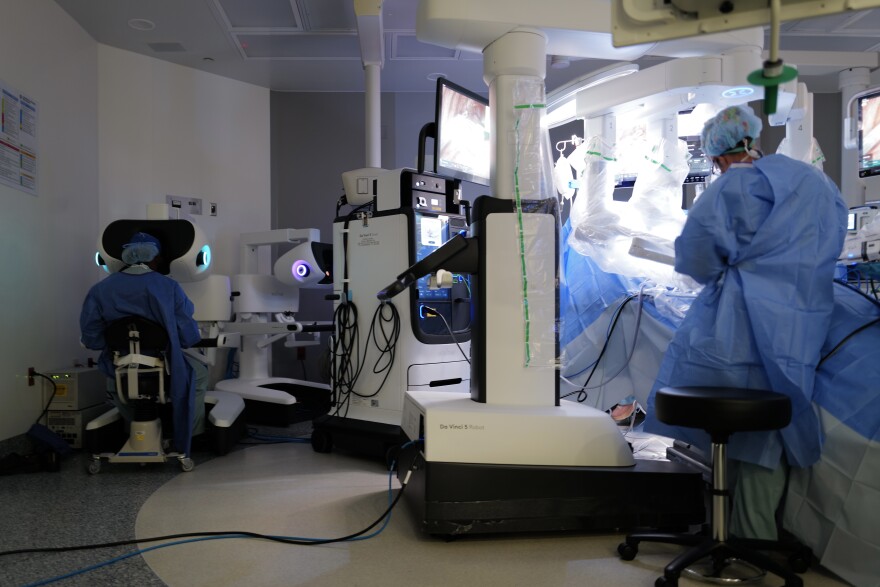 The main body of the Da Vinci 5 surgical robot stands beside the operating table, while surgeons monitor and control it from a nearby console.