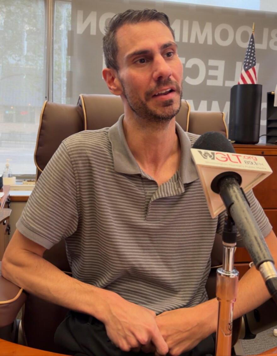 Man with a dark beard and wearing a striped brown shift sits behind a microphone with a flag that reads "WGLT.org 89.1 FM." A U.S. flag rests on a cabinet behind him.