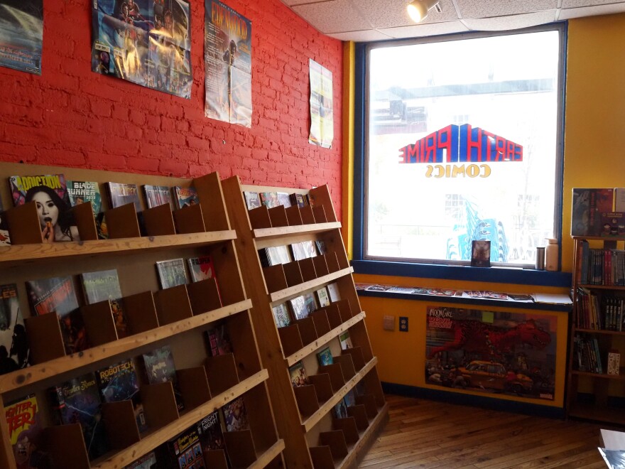 Light comes into a room from a window on a wall in front and to the right. Inside on the left is a red-painted wall with large wood bookshelves full of comic book covers. There are a few posters hanging above the shelves.