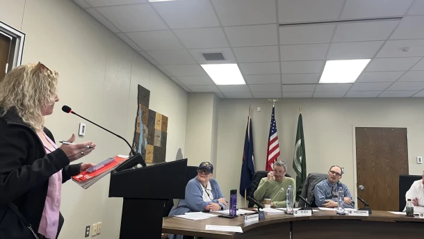 A person at a lectern addresses the Antrim County Commission. County Clerk Victoria Bishop, wearing a ballcap, is seated with three other people at a curved table facing the speaker.