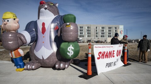 Three men can be seen standing outdoors on a corner of an intersection. They are standing behind a banner that reads "Shame on Port KC." At left is a large inflatable cat that is is smoking a cigar wearing a suite. It  has its paws wrapped around an inflatable person who looks like a construction worker in its right paw and a bag of money in its left paw.