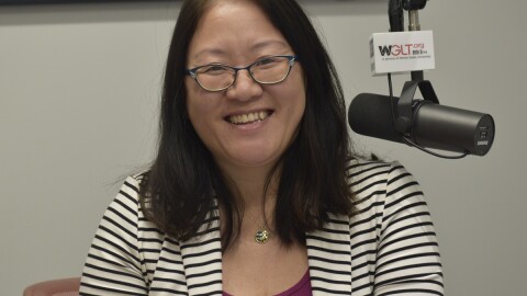 State Rep. Sharon Chung in a radio studio in a black-and-white striped suit jacket seated next to a microphone with the mic flag labeled 'WGLT.org 89.1 FM'