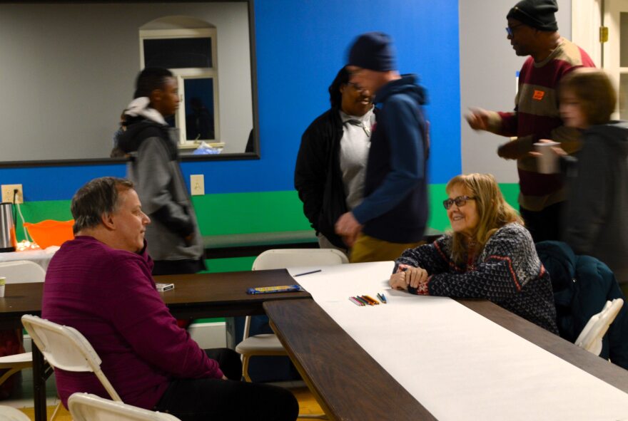 A man in a long sleeved purple sweater sits at a table talking to a woman with long blond hair and glasses.
