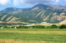 A green farm field in a mountain valley. There are some houses in the near background with green-covered mountain peaks rising up behind them.
