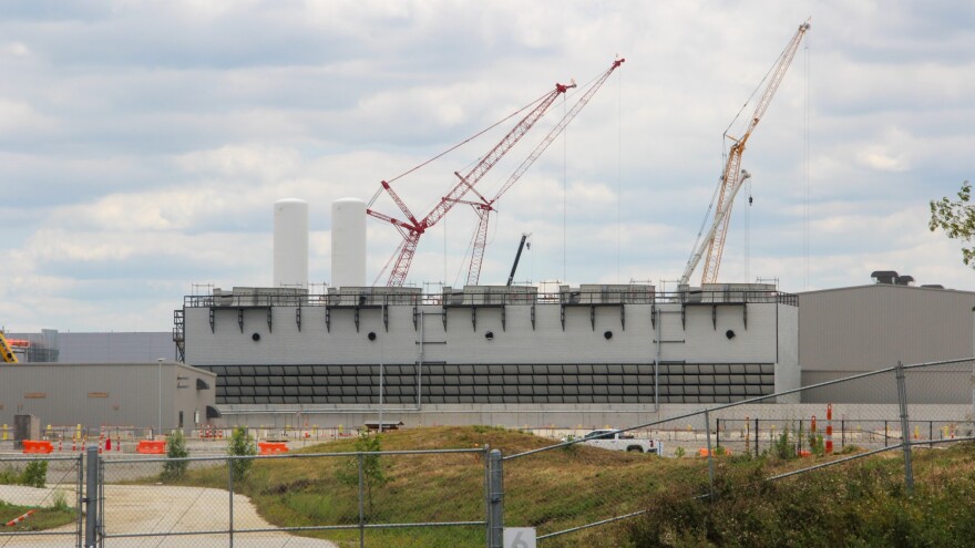 Tall cranes rise above a large, windowless building behind a chain fence. 