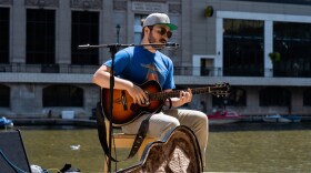 A man wearing a blue t-shirt, sunglasses and gray baseball cap plays an acoustic guitar and sings into a microphone while seated on a sidewalk next to a river in a downtown city setting.