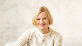 A woman with short blonde hair wears a white sweater and blue jeans and sits on a white couch. She crosses her hands and smiles at the camera. 