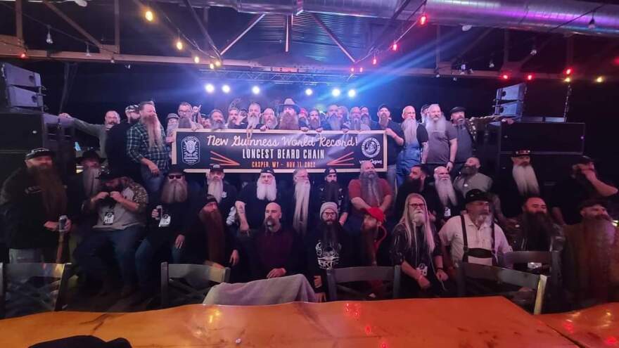 A group of bearded men gather around a banner proclaiming "New Guinness World Record! Longest Beard Chain Casper, WY - Nov. 11, 2022"