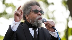 A man in sunglasses speaks into a mic held in one hand as he points at a high angle with the other. He's wearing a black suit jacket with a matching tie, along with a long sleeve white dress shirt with gold colored cufflinks. The background is heavily blurred, highlighting the speaker. 