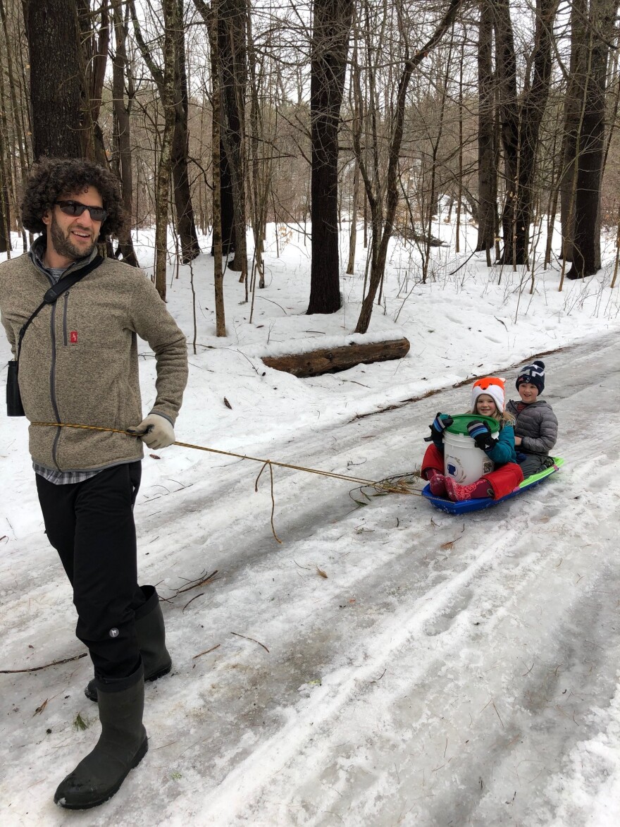 A man pulls a sled with a bucket of sap and 2 kids on a wintry road