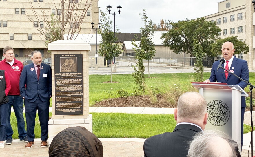 A man in a suit speaks at a podium with the Bradley University logo while two men stand next to a memorial plaque titled “The Goldin Gateway.”