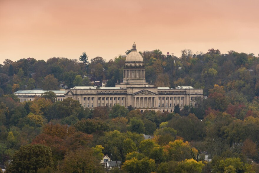 The Kentucky Capitol complex is shown from a high, far-away viewpoint, surrounded by trees and beneath a reddish sky.