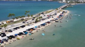 A stretch of road going through a blue body of water. On either side are short sand banks and dozens of parked vehicles and swimmers.