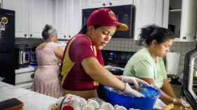 Three women cook in a kitchen; One is wearing a baseball cap that is maroon and yellow with a matching shirt. Another woman is wearing a pink and white dress and another is wearing a light green shirt. 