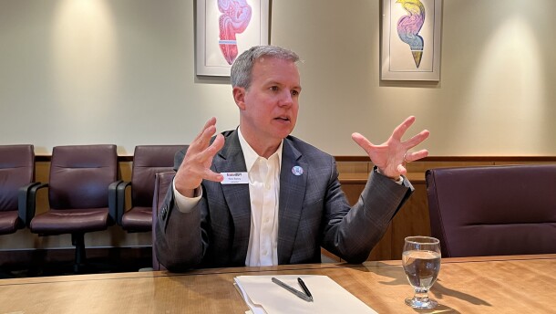 A middle aged man with gray hair sits at a conference table. He is talking and gesturing with his hands. 