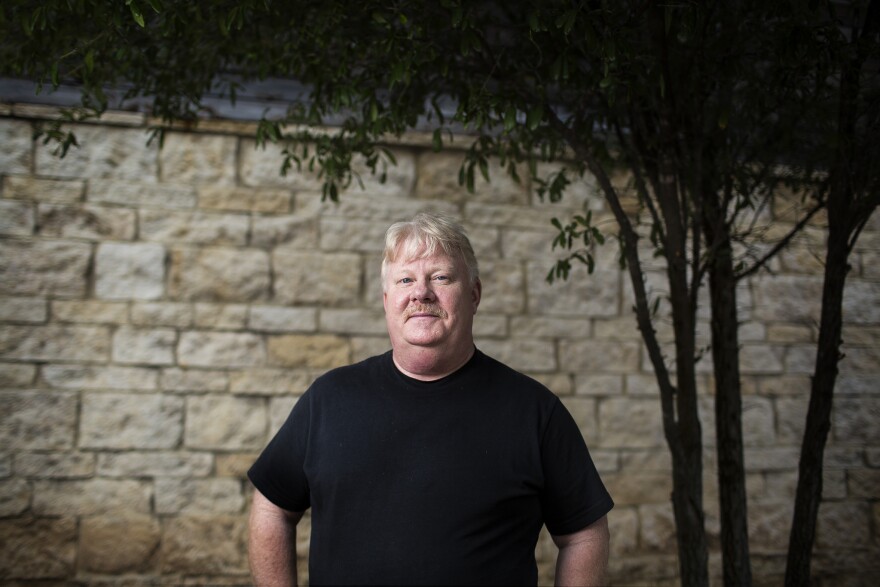 A man, Tracy Cluck, wearing a black shirt standing in front of a brick wall. 