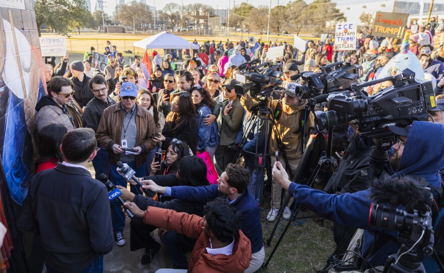 Reporters and television cameras, at right, point microphones to a group of people standing against a colorful wall. 