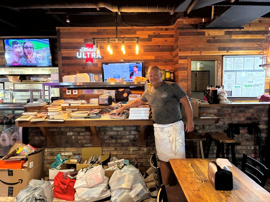 Alt text:

A man in a gray T-shirt and white apron stands smiling beside a large stack of secondhand books piled on a wooden bar counter inside a restaurant or pizzeria. More books and bagged donations fill boxes and shopping bags on the floor below. A neon Michelob Ultra sign, string lights, and exposed brick line the wall behind him, with a TV playing sports coverage and a community bulletin board visible in the background.