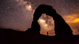 A person standing under the silhouette of the Delicate Arch rock formation at night. The sky is completely lit up with stars and galaxies.