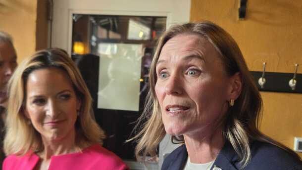 In the foreground, Winnie Brinks wears a navy blazer, American flag pin, and gold necklace. Jocelyn Benson stands beside her in a bright pink dress, smiling and looking toward Brinks. The two women are in a warmly lit room.