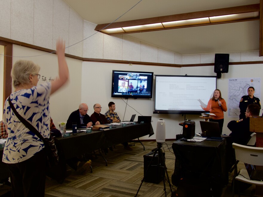 A woman standing at the back of a room raises her hand as another woman at the front speaks into a microphone.
