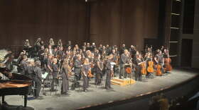 An orchestra all wearing black facing the audience on a grey stage.