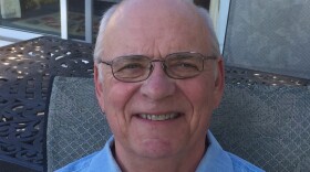 A smiling man with short white hair, wearing glasses and a blue collared shirt