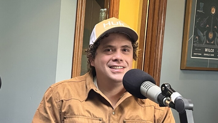 A man in a tan jacket and yellow baseball cap sits behind a microphone in the KAXE studio.