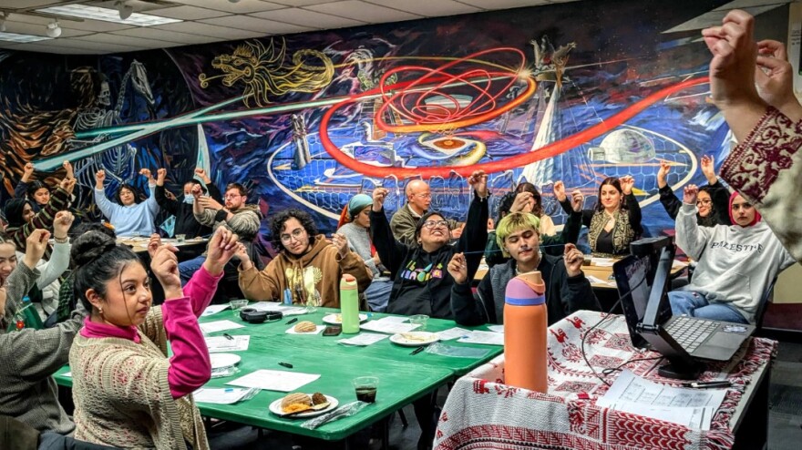 More than a dozen students hold thread in the air and look forward toward an instructor in a classroom filled with large tables and vibrant art on the walls.