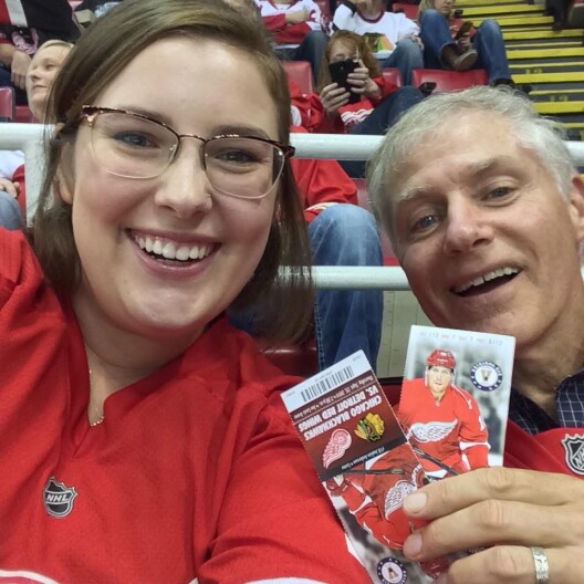 A selfie shows a young woman and an older man smiling in a stadium setting. Both individuals are looking directly at the camera, wearing Red Wings apparel. The woman wears glasses and has brown hair, while the man has short grey hair. The man holds up two printed hockey game tickets, showcasing logos and an image of a player. The ticket text reads "Chicago Blackhawks vs. Detroit Red Wings." The background features stadium seating with other spectators visible.