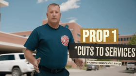 A men wearing a shirt with a logo of a fire department stands in front of a building. Words are superimposed over the image reading, "Prop 1 'Cuts to services' Wyoming Tribune Eagle, 7/31/26."