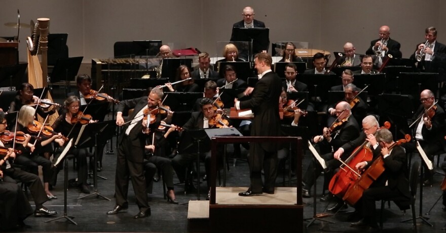 An orchestra plays and a violinist stands for his solo as the conductor looks at him from a small platform