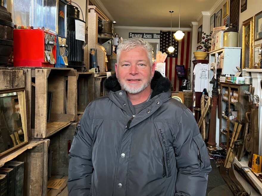 Lee Dowdy smiles for a portrait. He's bundled up in a black coat and standing amid shelves inside Fieldview Acres Mercantile.