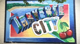 The image shows a post-card style mural paint on a beige brick wall that says “Greetings from Traverse City” in giant 3D block letters. The lettersT are filled with detailed illustrations of local features, including a lighthouse, sailboats, sand dunes, wine, and ski slopes. In the top right corner a pair of giant glossy red cherries sit. 