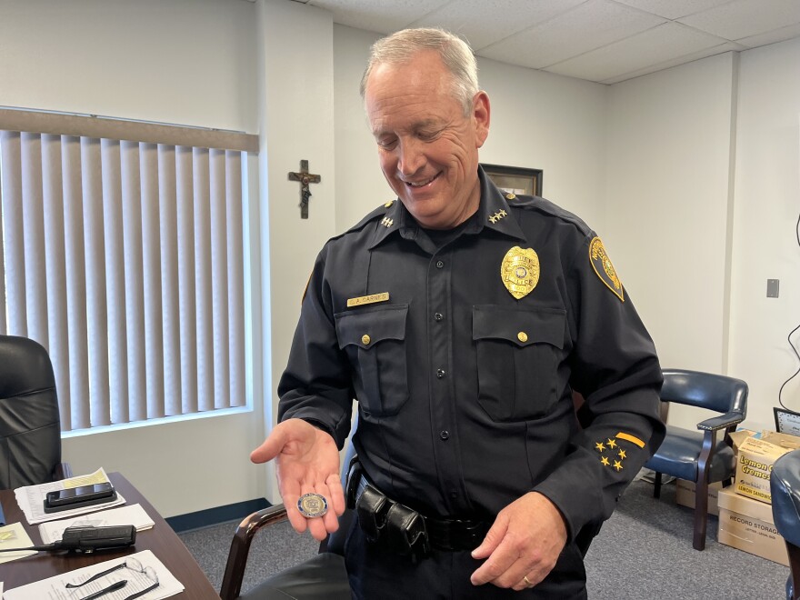 A man in a police uniform smiles as he looks at a coin in his hand. He's standing in an office with a cross hanging on the wall behind him and glasses and radio on the desk next to him.