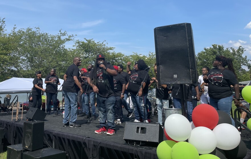 Under a blue sky a group of singers on stage, in black shirts, sing behind a man with a microphone in a baseball cap. 