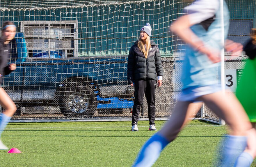 A woman with long blonde hair wearing a beanie, black pants and a black coat stands in front of a soccer goal speaking as blurred players run across the field.