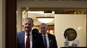 Two men in suits walk down a hallway.