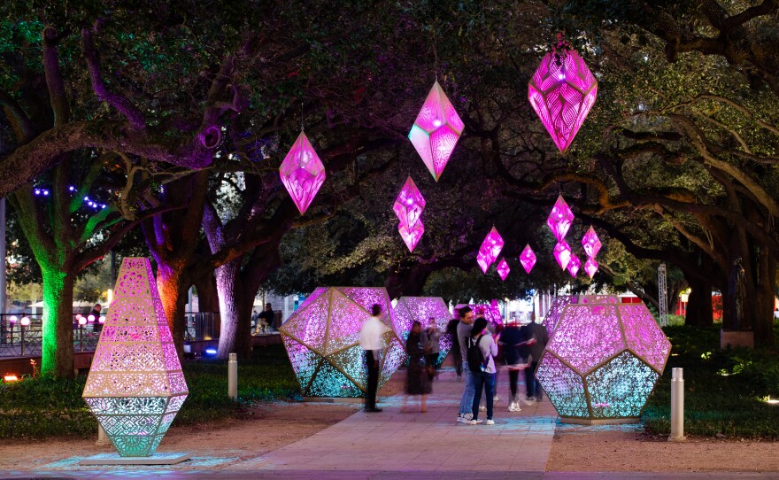Illuminated geometric shapes line a path through a tree-filled park at night.
