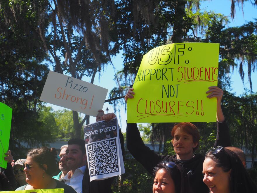 Standing in a park, people hold signs against closing Pizzo Elementary