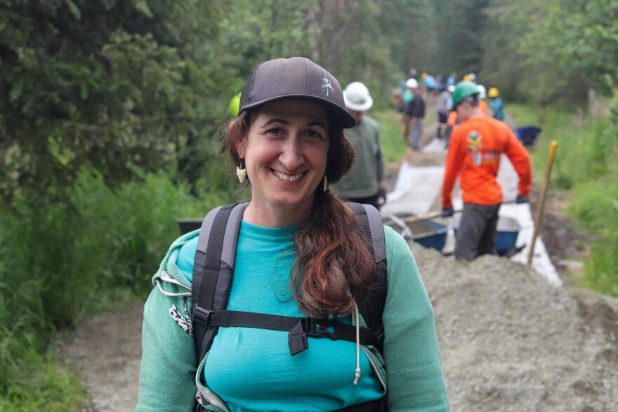 A woman with a baseball hat. Behind her there are people working on a trail, out of focus.