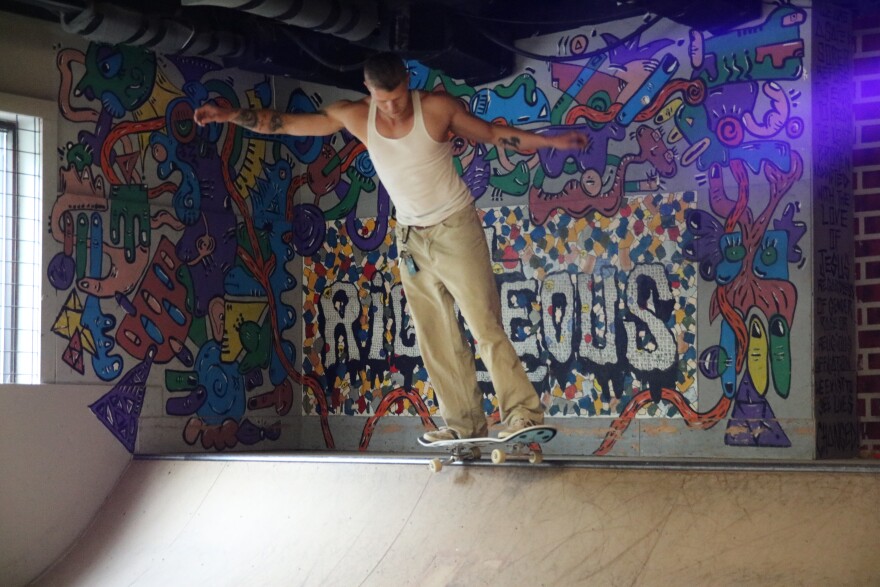 A guy in a white tank top and khaki pants balances on the edge of a wooden ramp on top of a black skateboard. Behind him is colorful graffiti that reads "Righteous" with the "t" being a cross.