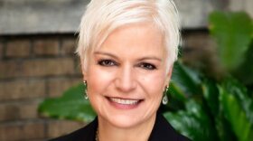 A headshot of Anne Constantino, wearing a black suit, white blouse and metal necklace.