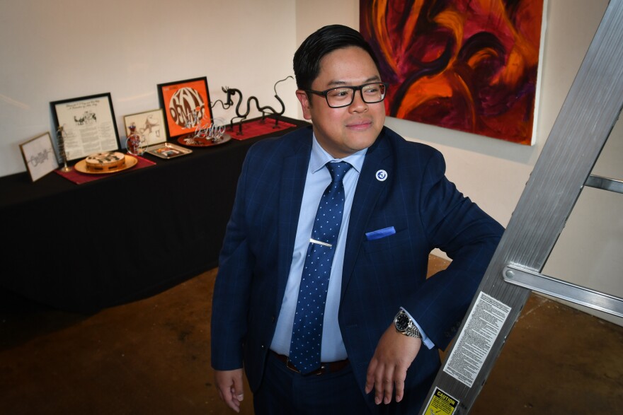 A Vietnamese man wearing a blue suit and tie leans on an aluminum ladder. Asian art sits on a table behind him and a colorful painting hangs on the wall.