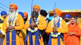 Five men holding swords walk in the procession behind devotees sweeping the ground with brooms, pouring water, and singing hymnals.