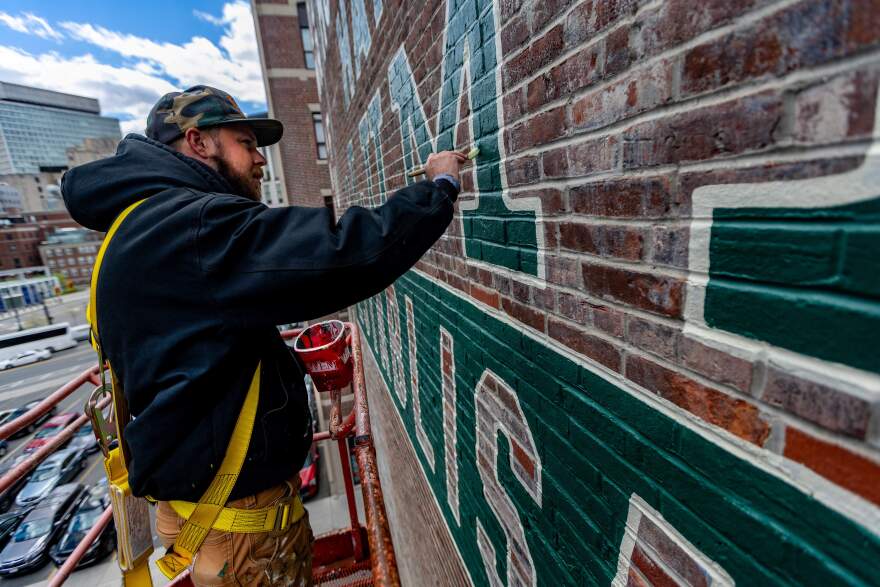 High above street level, Jack Williams applies paint to restore the Holt & Bugbee lumber company sign in the West End of Boston.