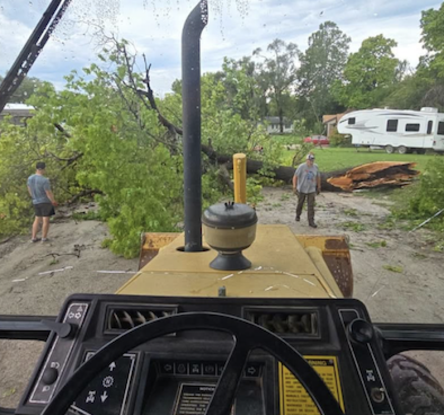 A resident uses a backhoe to remove fallen trees from a road near Howell after Thursday’s severe storms. Neighbors also brought chainsaws to help reopen the road.