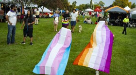 Two attendees of Boise Pride walk on the grass of Ann Morrison Park. One is wearing a Trans flag as a cape (Blue, pink, white) while the other is wearing a lesbian flag as a cape (pink, orange, yellow). 