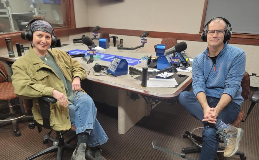 Two people wearing headphones sit at a table in a radio talk studio: a woman at left has a blue headscarf and is wearing a green jacket, green shirt, blue jeans and grey boots; a man at right has very short dark hair and is wearing glasses, a light blue quilted sweatshirt, blue jeans and brown boots.