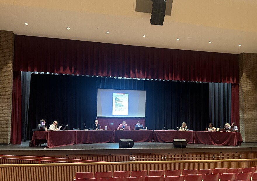 Nine people sit at five long tables lined up on a high school theater stage. A screen behind them shows the a document with the agenda.